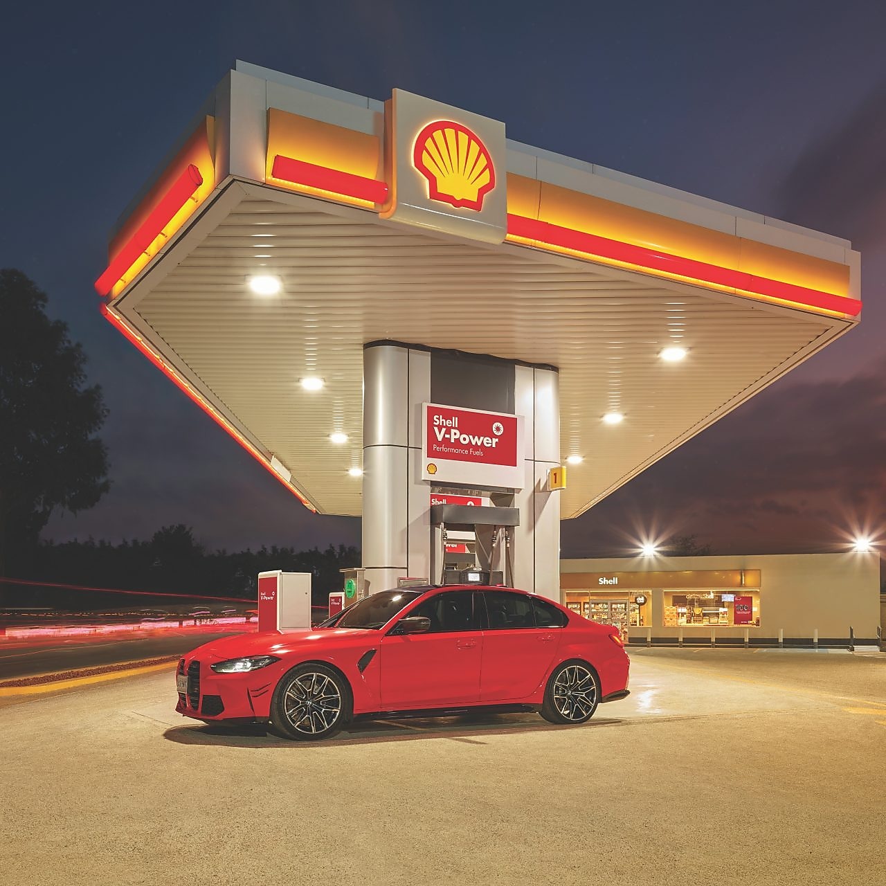 Červené auto stojace pred tankovacím stojanom čerpacej stanice Shell v nočných hodinách.