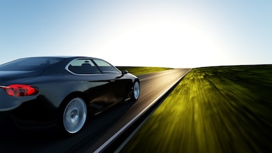 Čierne auto rýchlo idúce po asfaltovej ceste, rozmazaná tráva a obloha, jasné slnko na obzore.