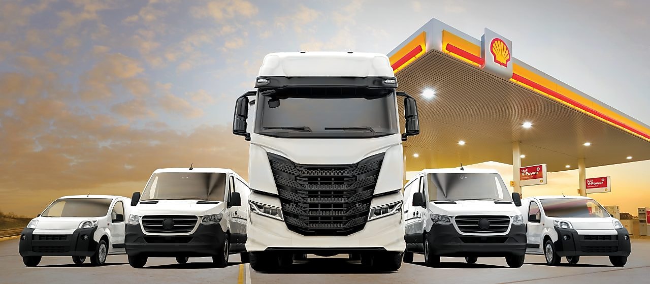 Biele úžitkové vozidlá, vrátane nákladných áut a dodávok, zaparkované symetricky pred stanicou Shell.