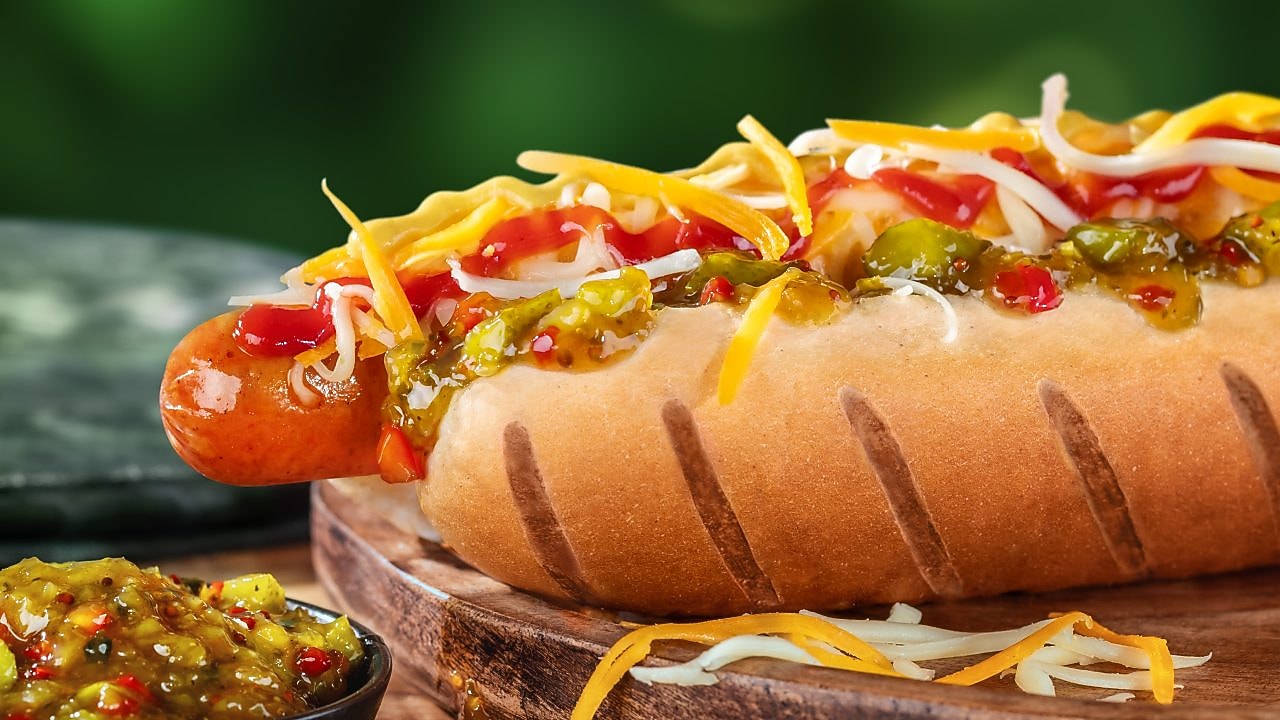 Foto hot dogu Mega s klobásou, karamelizovanou cibuľkou, kečupom, horčicou, syrom a uhorkovým relishom