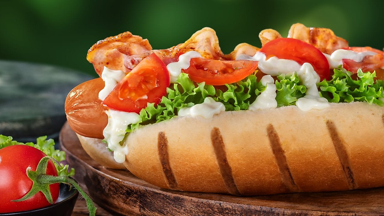 Foto hot dogu Anglcko so šalátom, paradajkou, majonézou a párkom so slaninou